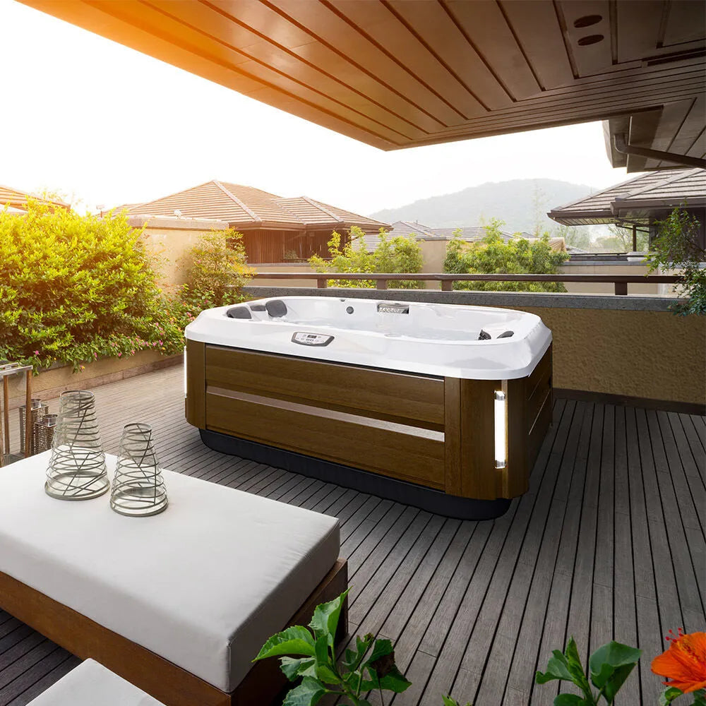 An outdoor J-315 COMFORT HOT TUB WITH LOUNGER FOR SMALL SPACES by Jacuzzi featuring PowerPro® jets is situated on a wooden deck under a covered area. The deck includes cushioned seating and decorative wire lanterns. Surrounding the deck are lush green plants, and additional housing structures can be seen in the background, enhancing relaxation with hydrotherapy benefits.