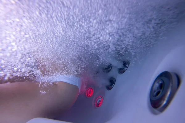 Underwater view of a person relaxing in a Jacuzzi - J-LXL Hot Tub by Jacuzzi. The water is bubbling vigorously from the jets, creating a frothy effect. The person is wearing white swimwear, and the red lights from the jets contribute to the relaxing ambiance, making it an ideal setting for hydrotherapy.