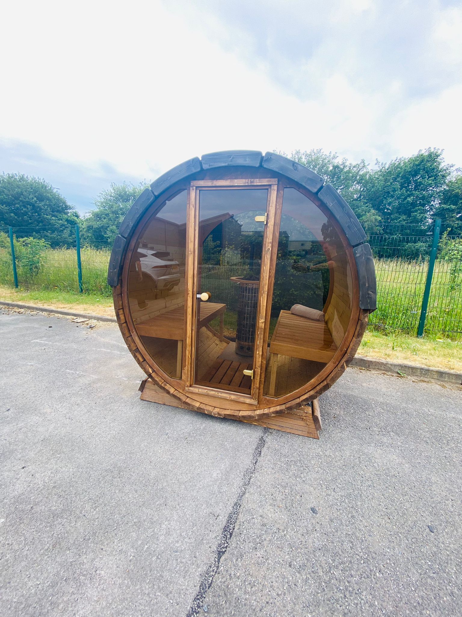 A cylindrical wooden outdoor pre-built Sunbeach Delux Moon Terrace 160 Sauna by Sunbeach Spas with a large glass door and windows, situated on a paved area with green trees and a fence in the background. The sauna features a bench inside, an electric Harvia heater, and a wooden roof with dark curved shingles.