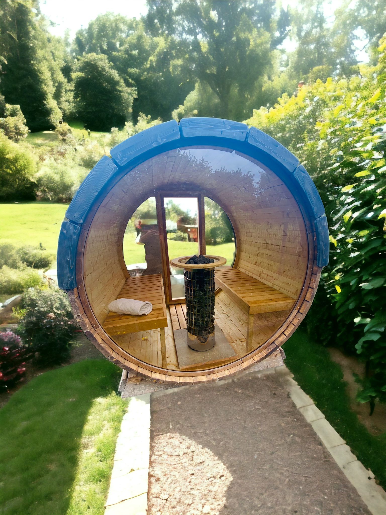 A Sunbeach Delux Moon Terrace 160 Sauna - Pre-Built with wooden bench seating and a central heating stove is situated on a garden path amid lush greenery. Through the glass door, a person can be seen standing, blending harmoniously with the serene outdoor environment, enjoying the warmth from an electric Harvia heater.