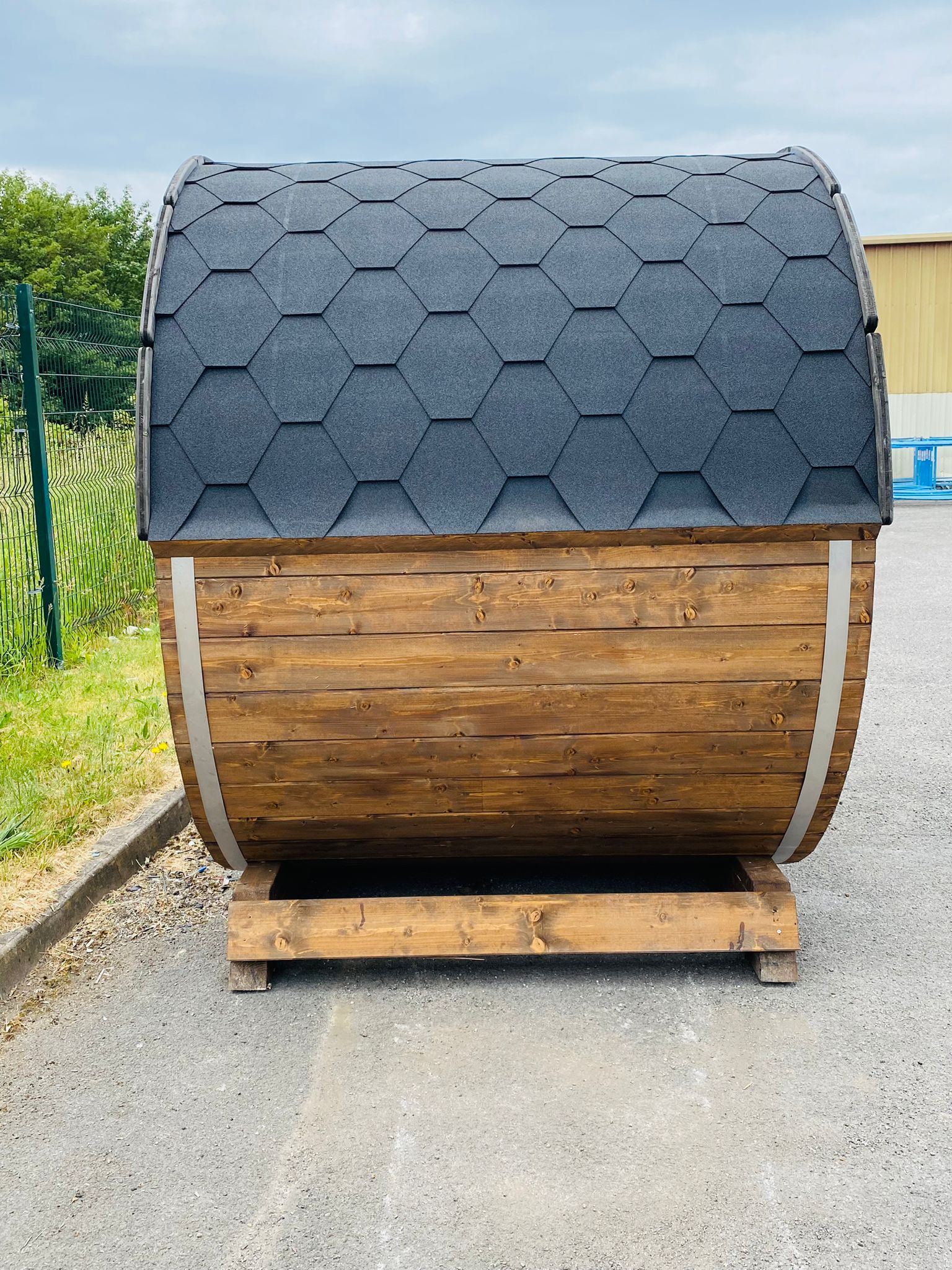 A small, wooden barrel-shaped **Sunbeach Delux Moon Terrace 160 Sauna - Pre-Built** with a hexagonal shingle roof is placed on a wooden pallet on a paved surface. It's situated outdoors near a green fence and some grass. Equipped with an electric Harvia heater, it's perfect for those cloudy days.
