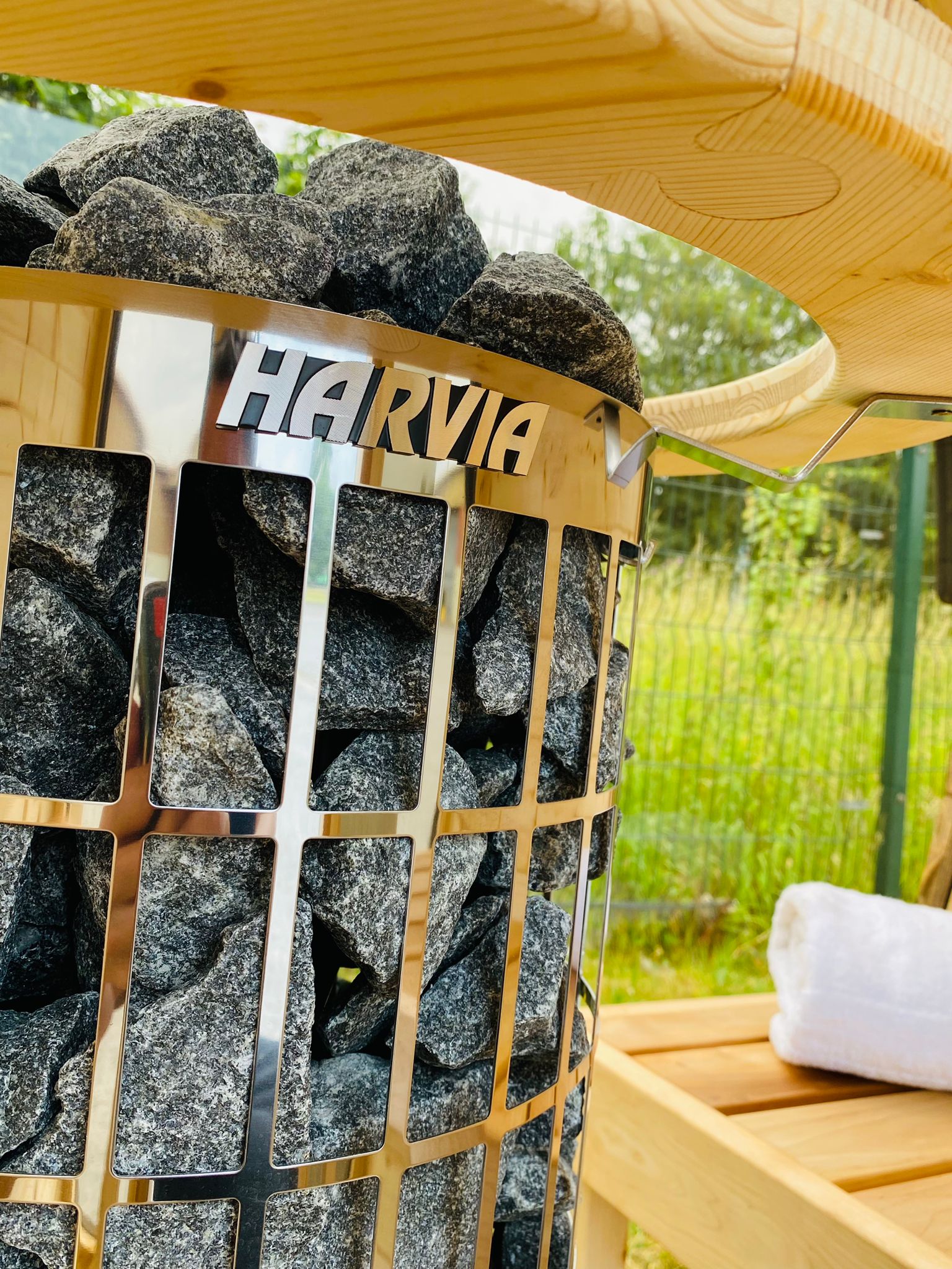 Close-up of an electric Sunbeach Delux Moon Terrace 160 Sauna - Pre-Built heater filled with dark sauna stones. The metallic heater features the Sunbeach Spas logo prominently. In the background, a rolled white towel is placed on a wooden surface, with a natural outdoor setting visible beyond. Ideal addition to any pre-built sauna.