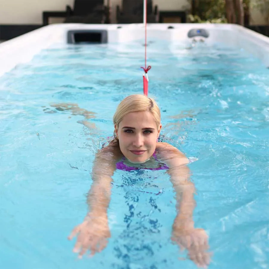 A person with blonde hair swims in a clear blue pool, attached to an Oasis Riptide - Easy Life Pro 8.0 with Inverter Swim Spa from Oasis Spas. The swimmer is seen facing the camera, with outstretched arms and a focused expression. The background shows poolside furniture and greenery.