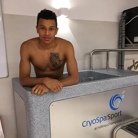 A shirtless man with a chest tattoo stands in a CryoSpa Sport - Cold Water Ice Bath. The CET CryoSpas logo and the hashtag “#chilling4champions” are visible on the side of the tub. He appears relaxed while undergoing athlete recovery ice bath therapy.