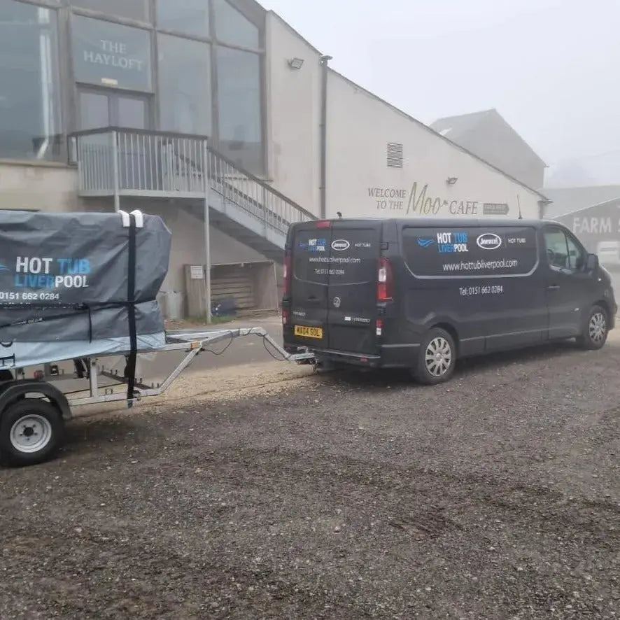 A van labeled "Hot Tub Relocation Services - Hot Tub Liverpool" with a phone number and website, parked outside a building labeled "The Hayloft" and "Moo Cafe" in North West England. The van is towing a covered hot tub on a trailer. The foggy, overcast setting adds an air of mystery to the scene.