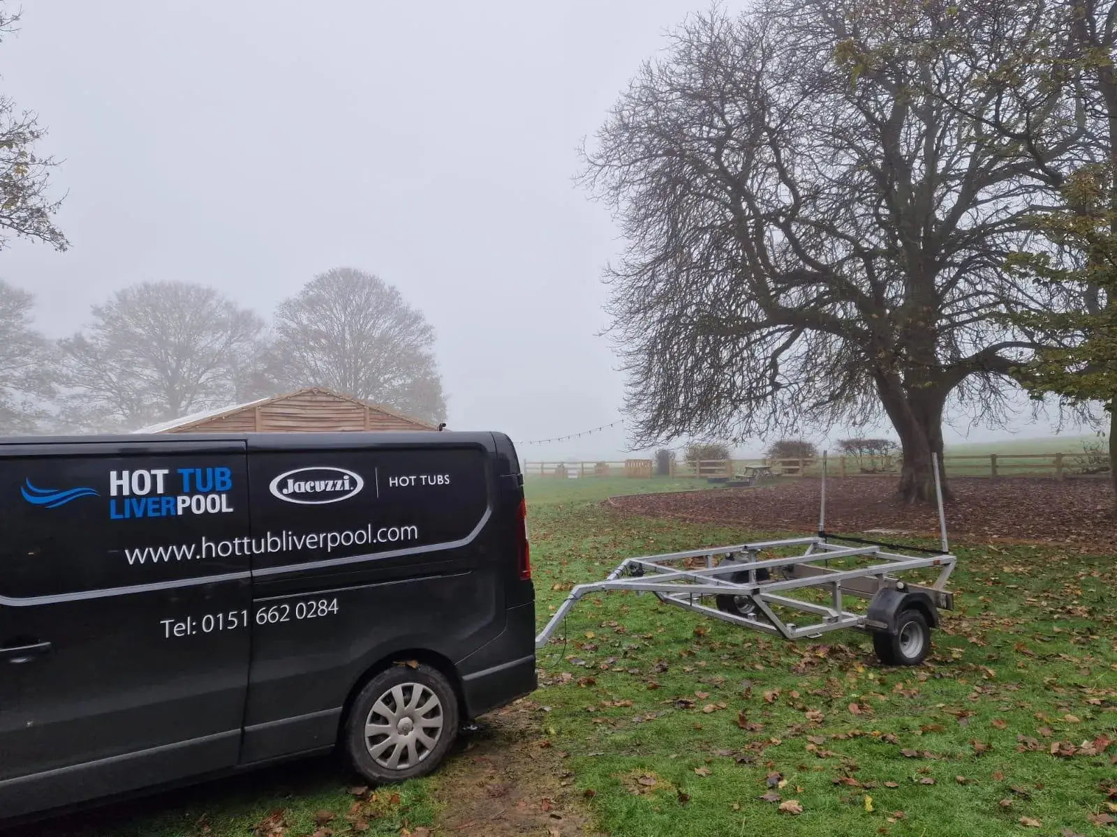 A black van with the branding "Hot Tub Liverpool" and a website URL "www.hottubliverpool.com" is parked on a foggy rural property in North West England. A tree with autumn leaves, a wooden shed, and an empty trailer for Hot Tub Relocation Services - Hot Tub Liverpool attached to the van can be seen in the background.