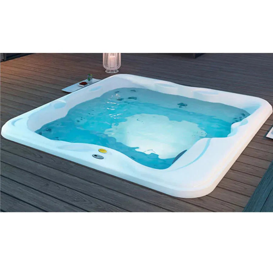 A modern, white square luxurious Jacuzzi Lodge+L Holiday Let Hot Tub filled with crystal-clear blue water, set flush with a wooden deck. The deck is empty except for a small table with a plant and a lantern providing soft lighting. The Jacuzzi hot tub features built-in seating, multiple jets, and an advanced water purification system.