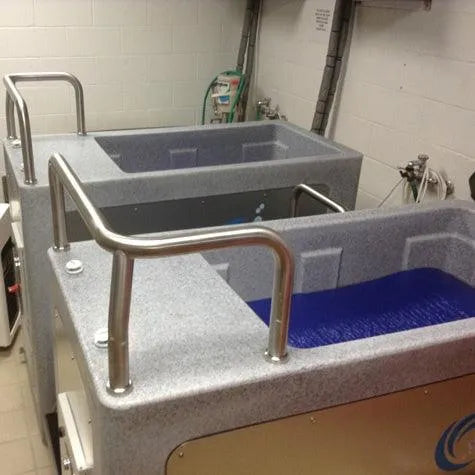 Two large therapy tubs with handrails in a treatment room designed for athlete recovery. One tub is filled with blue water, possibly indicating an ice bath, and the other is empty. These Team CryoSpa Sport Ice Bath tubs from CET CryoSpas are made of gray material with step access. Various pipes and hoses are visible in the background, attached to the wall.