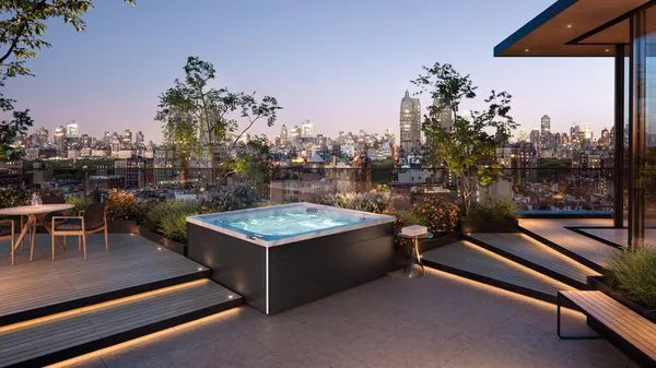 A rooftop terrace at sunset featuring a Jacuzzi - J-LXL Hot Tub with hydrotherapy surrounded by modern decking, lush plants, and seating areas. The terrace overlooks an urban cityscape with tall buildings and trees, illuminated by glowing city lights in the distance.