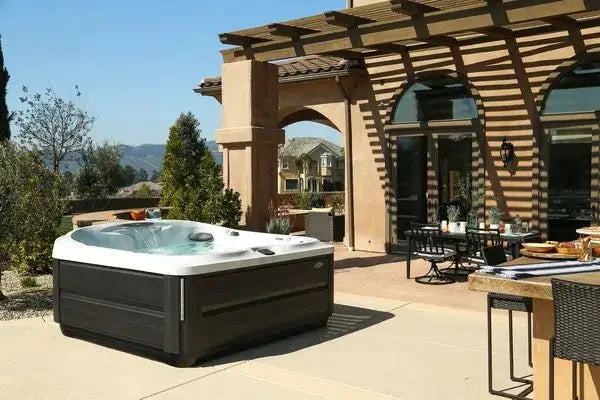 An outdoor patio features a Jacuzzi J-495™ DESIGNER ENTERTAINER'S HOT TUB WITH INFRARED & RED LIGHT THERAPY - NEW MODEL and a dining area with chairs and a table set for a meal. The space is shaded by a pergola attached to a stucco house with arched windows. In the background, trees and distant hills stretch under a clear blue sky.