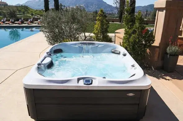 A luxurious Jacuzzi J-495™ DESIGNER ENTERTAINER'S HOT TUB WITH INFRARED & RED LIGHT THERAPY - NEW MODEL with bubbling water set on a patio beside a swimming pool. The background features a lush garden with manicured greenery, lounge chairs, and scenic mountains under a clear sky, promising an unparalleled hydromassage experience with PowerPro jets.