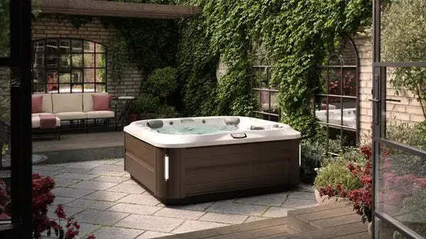 A cozy backyard patio features a J-335 COMFORT HOT TUB WITH OPEN SEATING by Jacuzzi, surrounded by green ivy-covered brick walls. A comfortable seating area with a cushioned sofa and matching chairs is visible on the left, bordered by vibrant red and pink flowers.
