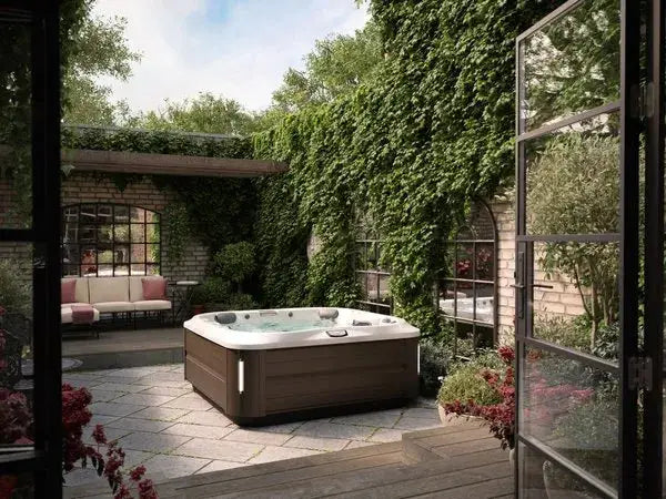 A cozy patio features a J-335 Comfort Hot Tub with Open Seating by Jacuzzi, surrounded by lush green ivy climbing the walls. There is a comfortable outdoor sofa with red and white cushions, a small table, and blooming potted plants. The scene is viewed through open glass doors, creating an inviting atmosphere.