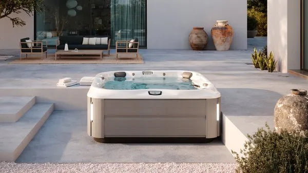 A modern, outdoor patio area featuring a luxurious Jacuzzi J-375 COMFORT HOT TUB WITH LARGEST LOUNGE SEAT NEW with advanced hydromassage technology. The patio is adorned with minimalist furniture, large clay pots, and greenery. In the background, there's a glass-walled room with additional seating, blending indoor and outdoor relaxation spaces.