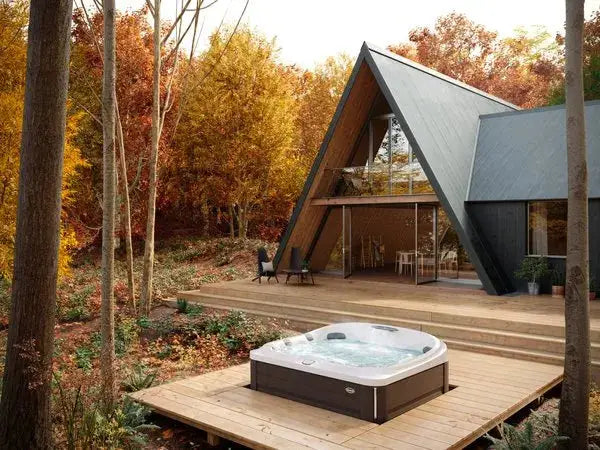 A modern A-frame cabin with large windows nestled in an autumn forest. The cabin features a spacious deck with a J-445™ OPEN SEATED HOT TUB WITH INFRARED & RED LIGHT THERAPY by Jacuzzi in the foreground and outdoor seating. The surrounding trees display vibrant fall foliage, creating a cozy, natural atmosphere.
