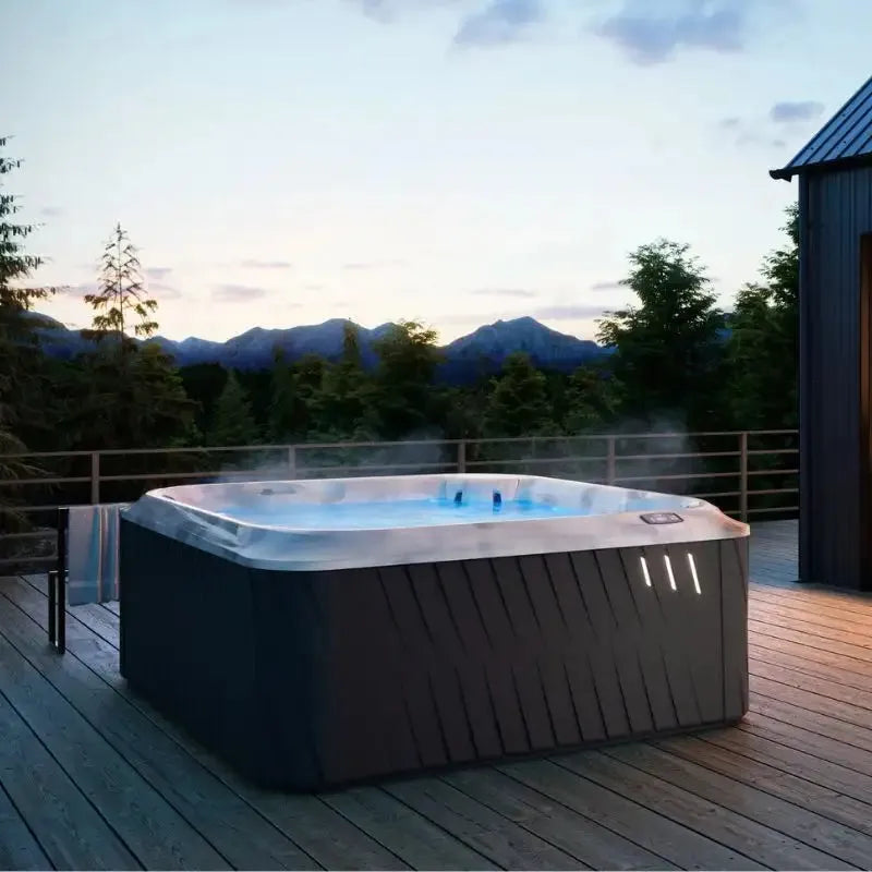 A Jacuzzi J-275 CLASSIC LARGE HOT TUB WITH LOUNGE SEAT - NEW MODEL sits on a wooden deck in an outdoor setting. Steam rises from the warm water, and in the background, there are trees and a view of mountains under a peaceful twilight sky. The atmosphere suggests a tranquil and relaxing environment.