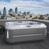 A petite rooftop hot tub with a sleek, modern design sits on a terrace overlooking a city skyline. Tall buildings and skyscrapers rise in the background under a partly cloudy sky. Equipped with PowerPro® jets, the J-315 COMFORT HOT TUB WITH LOUNGER FOR SMALL SPACES by Jacuzzi offers remarkable hydrotherapy benefits and various control features.