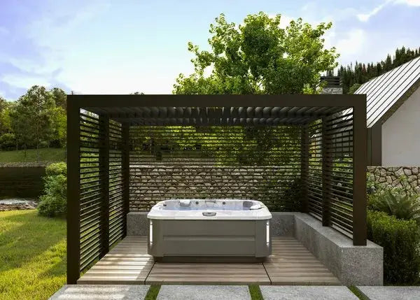 A modern outdoor hot tub featuring the Jacuzzi® J-335 Comfort Hot Tub with Open Seating sits under a sleek pergola with horizontal slats, surrounded by lush greenery. The pergola provides partial shade, and there is a stone pathway leading to the hot tub. In the background, a house with a pitched roof is partially visible.