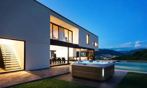 A modern two-story house with large windows is lit up at dusk. The backyard features a wooden hot tub equipped with the J-335 COMFORT HOT TUB WITH OPEN SEATING by Jacuzzi and an illuminated swimming pool. In the background, there are mountains and a partially cloudy sky, with a scenic city view in the distance.