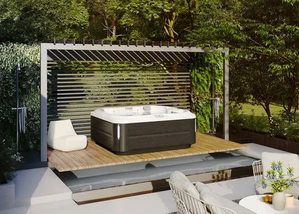 A modern outdoor patio featuring a Jacuzzi J-355 HOT TUB WITH COMFORT LOUNGE SEATING & COOL DOWN SEAT shaded by a slatted pergola. The hot tub, equipped with ProClear filtration system and hydrotherapy jets, is set on a wooden deck with an adjacent white lounge chair. Lush greenery surrounds the patio, and comfortable outdoor seating is visible in the foreground.