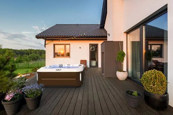 A modern house with a wooden deck featuring a Jacuzzi J-355 Hot Tub with Comfort Lounge Seating & Cool Down Seat, equipped with hydrotherapy jets, surrounded by potted plants. The deck offers a scenic view of a green landscape. Large sliding glass doors lead into the house, and string lights hang along the roofline. The sky is clear with a few clouds.
