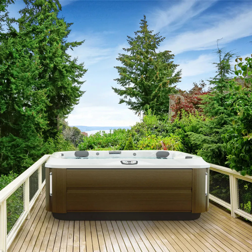 A luxurious J-375 COMFORT HOT TUB WITH LARGEST LOUNGE SEAT NEW by Jacuzzi sits on a wooden deck surrounded by lush greenery and tall trees, with a view of the sky and distant mountains. The scene is calm and inviting, ideal for relaxation, enhanced by advanced hydromassage technology.