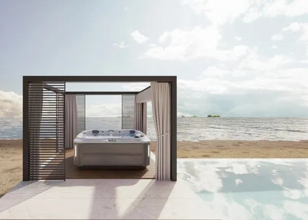 A modern outdoor spa setup by the beach features a J-375 COMFORT HOT TUB WITH LARGEST LOUNGE SEAT NEW by Jacuzzi under a sleek, open gazebo with wooden slats on the sides. Cream-colored curtains hang down partially, and a calm ocean with a partly cloudy sky is visible in the background, showcasing advanced hydromassage technology for ultimate relaxation.