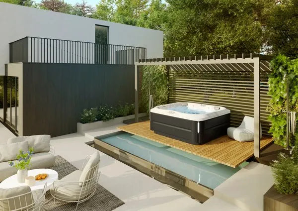 A modern backyard features a Jacuzzi J-375 COMFORT HOT TUB WITH LARGEST LOUNGE SEAT NEW with advanced hydromassage technology on a wooden deck surrounded by greenery. A pergola provides partial shade. Adjacent is a shallow, narrow pool. White outdoor furniture with a round table and chairs sits on a patio, near the contemporary house.