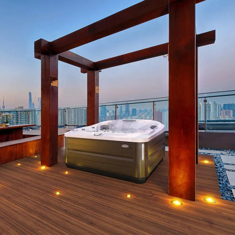 A rooftop deck with a modern J-445™ Open Seated Hot Tub with Infrared & Red Light Therapy under a wooden pergola. The deck features recessed lighting in the wooden floorboards. In the background, there's a clear glass railing providing a view of a city skyline at dusk. Indulge in ultimate relaxation with Jacuzzi PowerPro jets offering premium hydrotherapy.