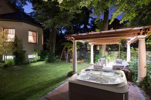 A nighttime outdoor scene features a Jacuzzi J-445™ Open Seated Hot Tub with Infrared & Red Light Therapy on a wooden deck under a pergola adorned with hanging plants. Nearby is a cozy seating area with cushioned chairs and a small table, surrounded by lush greenery. A lit house and swing set are visible in the background.
