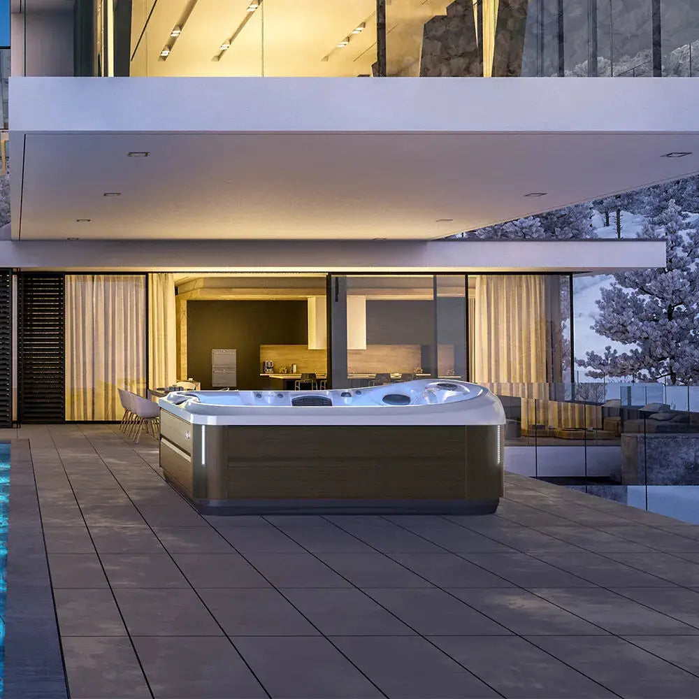 A modern outdoor patio at dusk with a large Jacuzzi J-495™ DESIGNER ENTERTAINER'S HOT TUB WITH INFRARED & RED LIGHT THERAPY - NEW MODEL in the foreground. The patio features sleek, minimalist design elements and a part of a contemporary building with large glass windows and a lit interior. Trees are visible in the background, promising an unparalleled hydromassage experience.