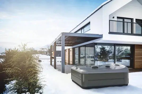 A modern, two-story house with large windows and a covered patio is seen in a snowy landscape. A Jacuzzi J-495™ DESIGNER ENTERTAINER'S HOT TUB WITH INFRARED & RED LIGHT THERAPY - NEW MODEL sits on the patio near some evergreen bushes, providing a relaxing contrast to the winter scenery. The sky is clear with the sun low on the horizon.