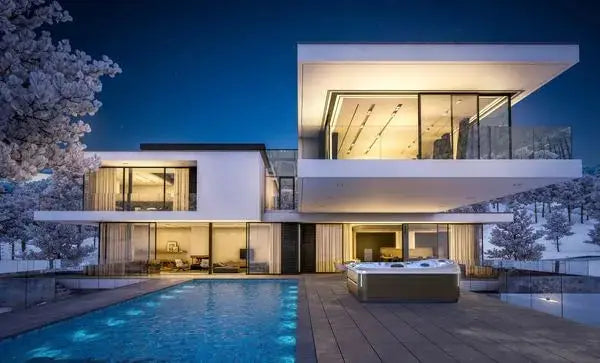 Modern two-story house at night with large glass windows, featuring illuminated interiors. The home has a spacious patio with a pool and a J-495™ DESIGNER ENTERTAINER'S HOT TUB WITH INFRARED & RED LIGHT THERAPY - NEW MODEL by Jacuzzi, surrounded by a snowy landscape with trees. The lighting creates a warm contrast against the cold, snowy environment for an unparalleled hydromassage experience.