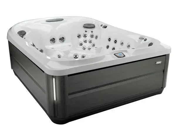 A modern, square-shaped Jacuzzi J-495™ DESIGNER ENTERTAINER'S HOT TUB WITH INFRARED & RED LIGHT THERAPY - NEW MODEL with sleek, dark grey exterior panels and a white interior boasts multiple PowerPro jets and built-in headrests. The hot tub features a digital control panel on the inner edge, offering a premium hydromassage experience.
