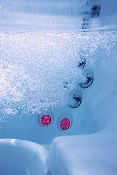 Underwater view of a Jacuzzi - J-LXL Hot Tub with multiple jets in operation, releasing streams of bubbles. Two red circular lights are illuminated just below the jets, casting a soft glow in the swirling water. The image captures a sense of relaxation and hydrotherapy in motion.