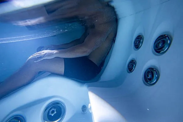 A person wearing black swim trunks is partially submerged in water, sitting against the side of a Jacuzzi - J-LXL Hot Tub with hydrotherapy jets visible. The underwater angle captures the reflection of the person on the water's surface.