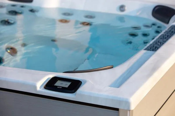 A close-up image of a white, rectangular Jacuzzi - J-LXL Hot Tub filled with clear water. The hot tub features built-in water jets for hydrotherapy, two black headrests, and a small digital control panel mounted on the side. The surface of the water reflects the surrounding light.