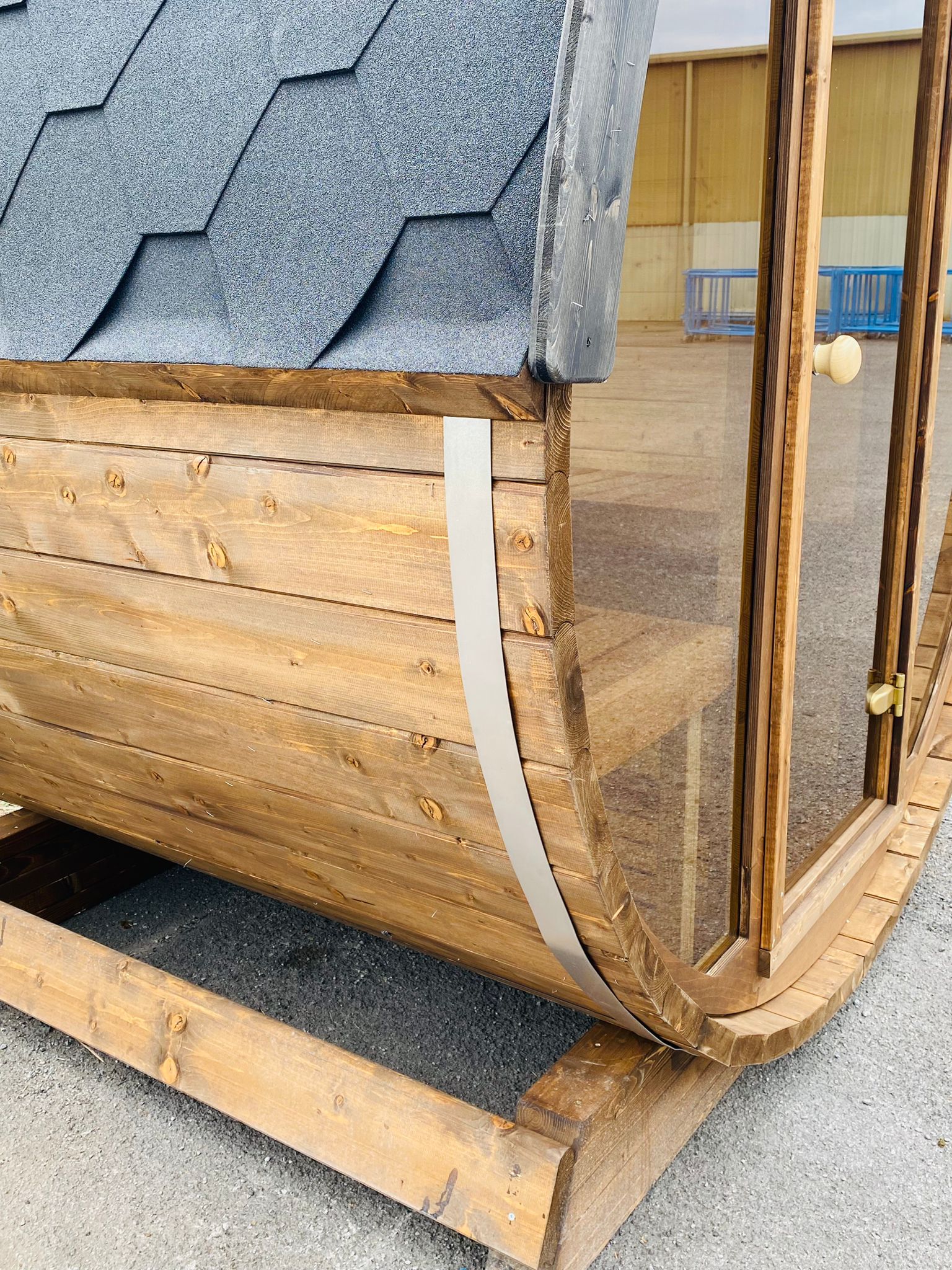 Close-up of a Sunbeach Spas Sunbeach Delux Moon Terrace 160 Sauna - Pre-Built with a wooden, curved design. The exterior features overlapping dark shingle roofing and light brown wood panels, with a metal band securing the corner. A partially visible indoor area with blue barriers and a sandy floor can be seen in the background.