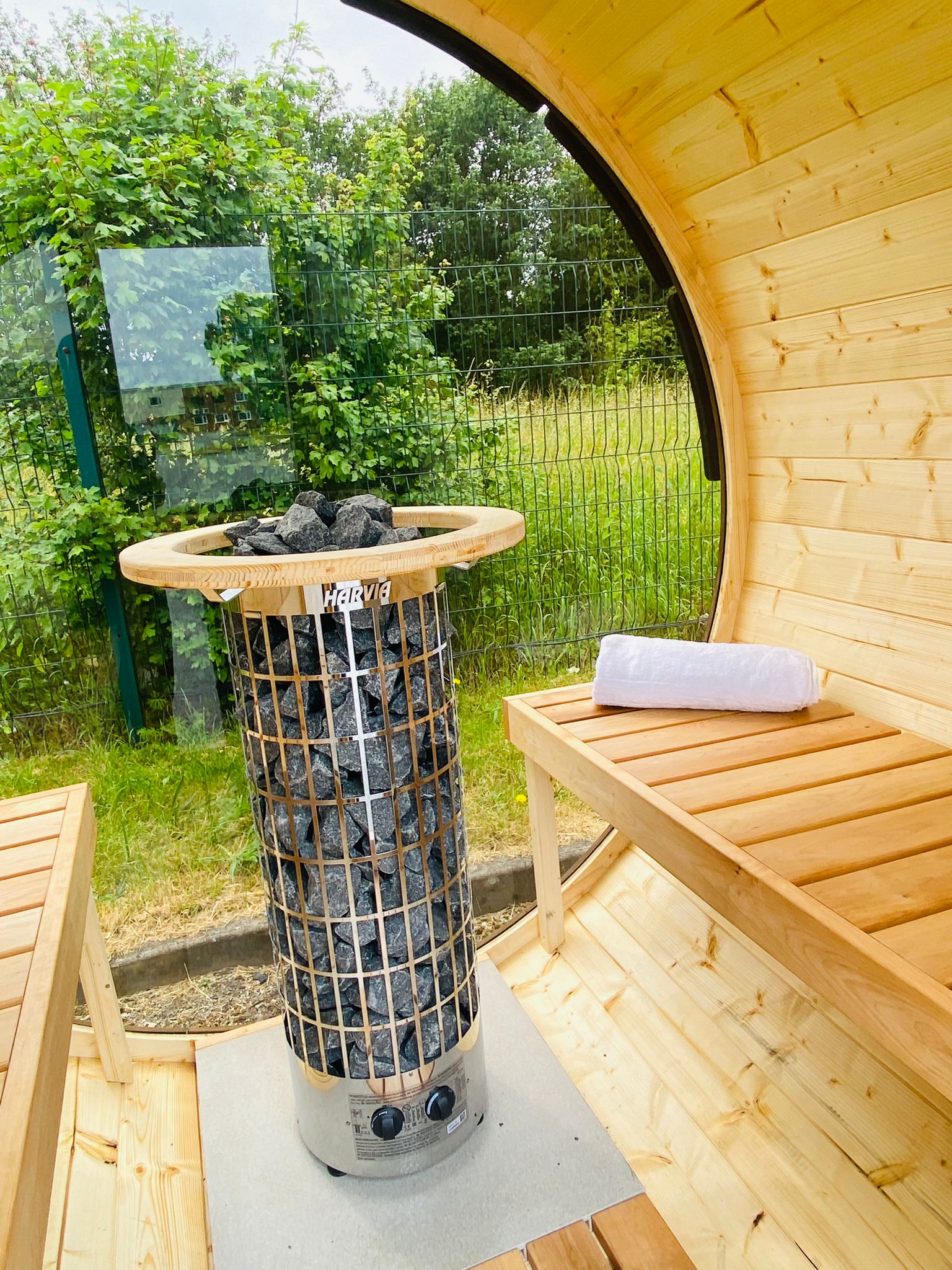 A cozy Sunbeach Delux Moon Terrace 160 Sauna - Pre-Built with a modern electric Harvia heater filled with rocks is in the center. Bench seating is on both sides with a towel rolled up on one bench. The sauna has glass windows offering a view of greenery and a fenced outdoor area.