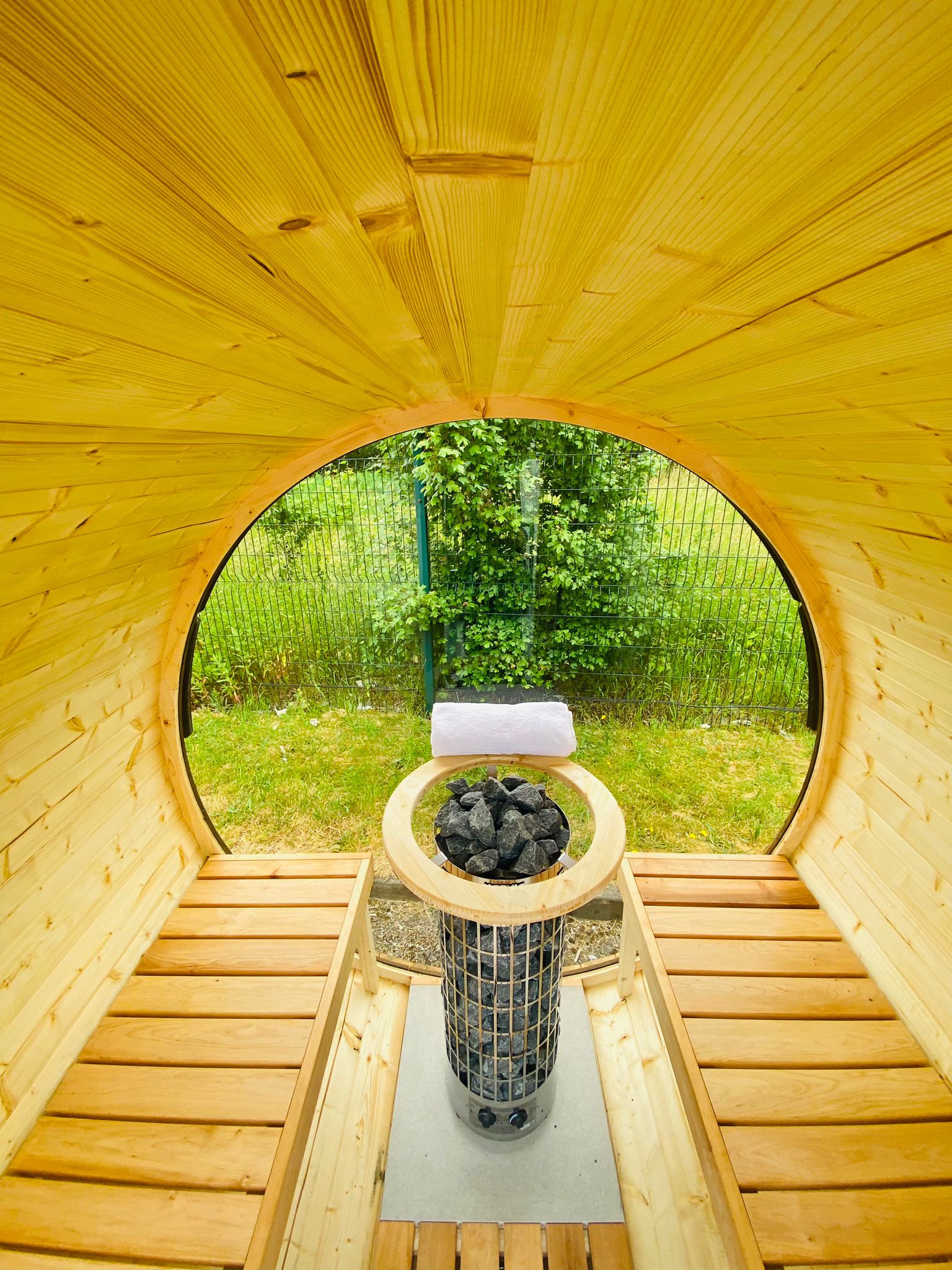 Interior view of a Sunbeach Delux Moon Terrace 160 Sauna - Pre-Built with wooden benches on both sides and an electric Sunbeach Spas heater filled with stones in the center. The room is illuminated by natural light through an open door, showcasing greenery outside.
