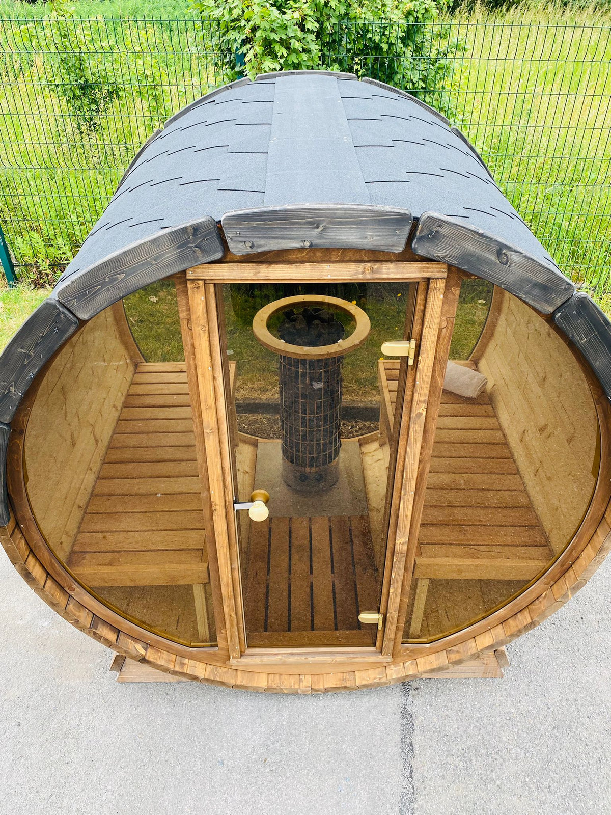 A Sunbeach Delux Moon Terrace 160 Sauna - Pre-Built by Sunbeach Spas made of wood with a clear glass door reveals two wooden benches and an electric Harvia heater inside. The dark-shingled roof adds a sleek touch, as the sauna sits on a concrete surface with grass and a fence in the background.