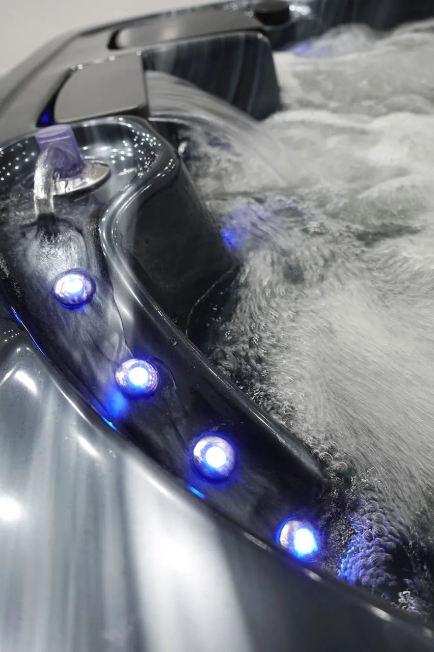Close-up of a modern Hot Tub Liverpool Atlanta - 2-3 Person Hot Tub with illuminated blue LED lights along the edge. Customizable jets create ripples and bubbles on the surface, and a stream of water pours into the tub from a small spout. The sleek, dark gray finish adds a touch of luxury to the setting.