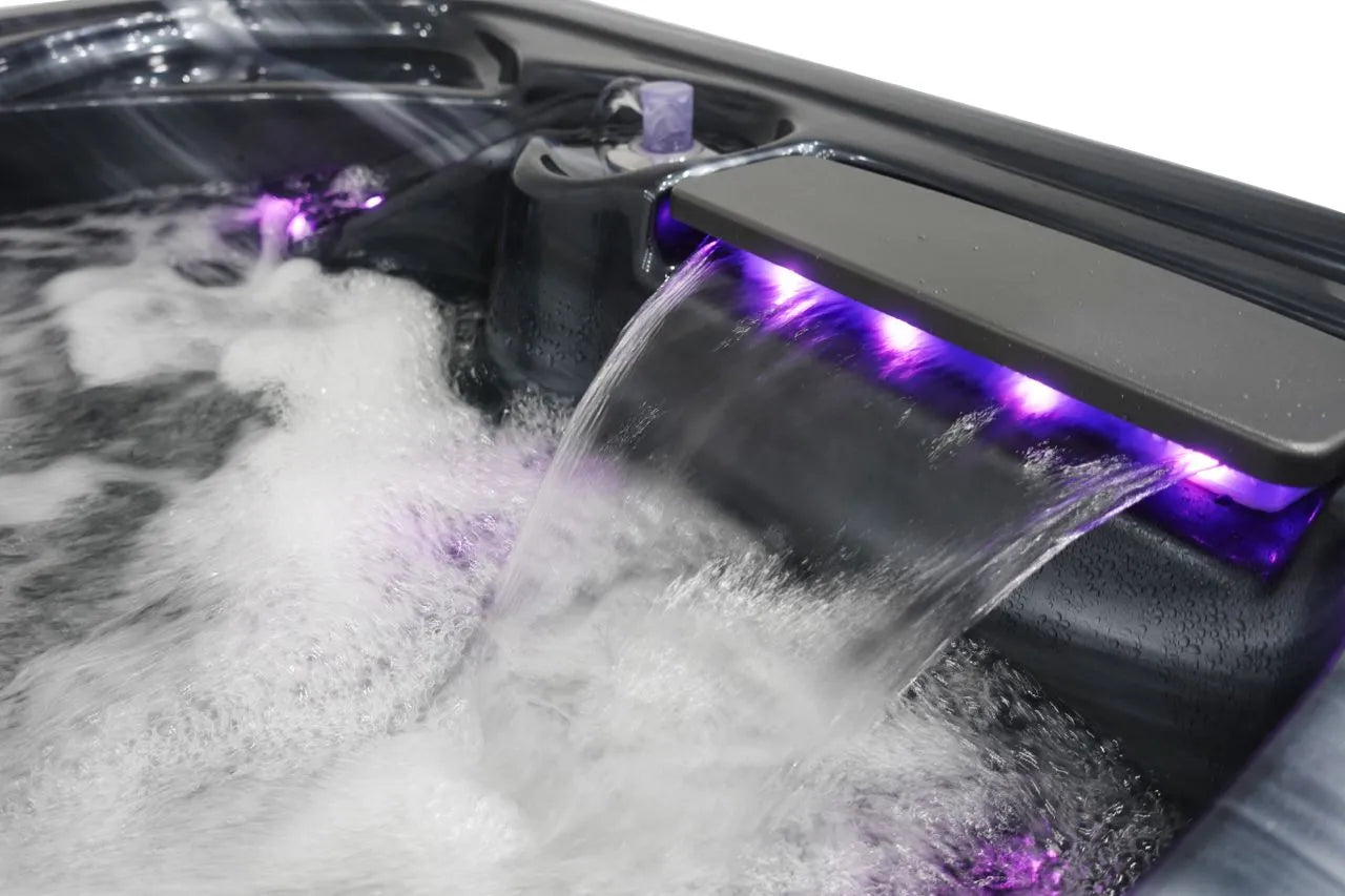 Close-up of an Atlanta - 2-3 Person Hot Tub by Hot Tub Liverpool with a cascading waterfall feature illuminated by purple LED lights. Bubbling water, controlled by the Balboa Control System, fills the tub, creating a relaxing and soothing atmosphere. The sleek, black interior of the hot tub contrasts with the bright purple glow of the lights.