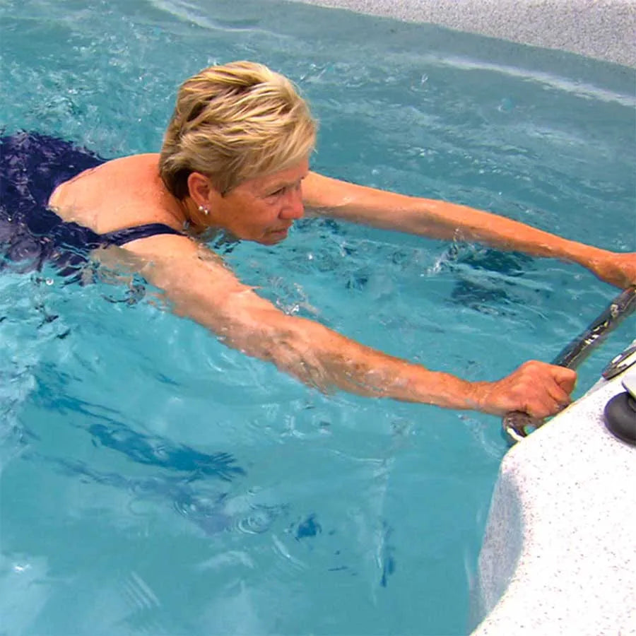 An older woman with short blonde hair wearing a blue swimsuit is swimming in a pool equipped with the Oasis Spas Oasis Riptide - Atlantis Pro Premium 4.4 Swim Pool with Inverter. She is holding onto a metal railing on the edge of the pool with both hands, seemingly taking a moment to rest or preparing to exit. The water is clear and blue.