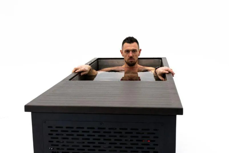 A man with short dark hair and a beard is submerged up to his shoulders in a rectangular Ice Tub. He is looking straight ahead with a neutral expression. The Chill Tubs - Professional affordable Ice Baths ideal for gym fanatics, designed by Chill Tubs, has a wooden rim and dark metal sides, and the background is plain white.