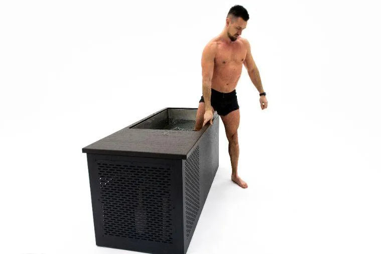 A shirtless man with black shorts and a tattoo on his left arm is standing beside a large black rectangular storage unit filled with water. He is preparing to immerse his right hand and part of his arm into the Chill Tubs - Professional affordable Ice Baths ideal for gym fanatics for cold water therapy. The background is plain white.