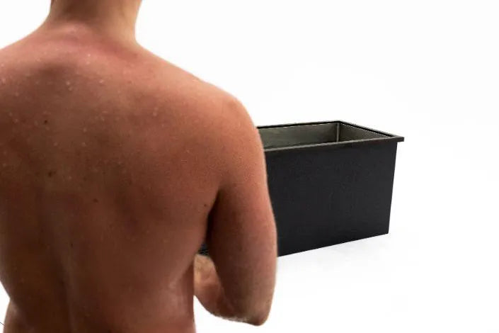 A shirtless person with visible back acne and shoulders stands in front of a black rectangular container against a white background. The container appears empty, resembling a Chill Tubs - Professional affordable Ice Baths ideal for gym fanatics, and the person is facing away from the camera.