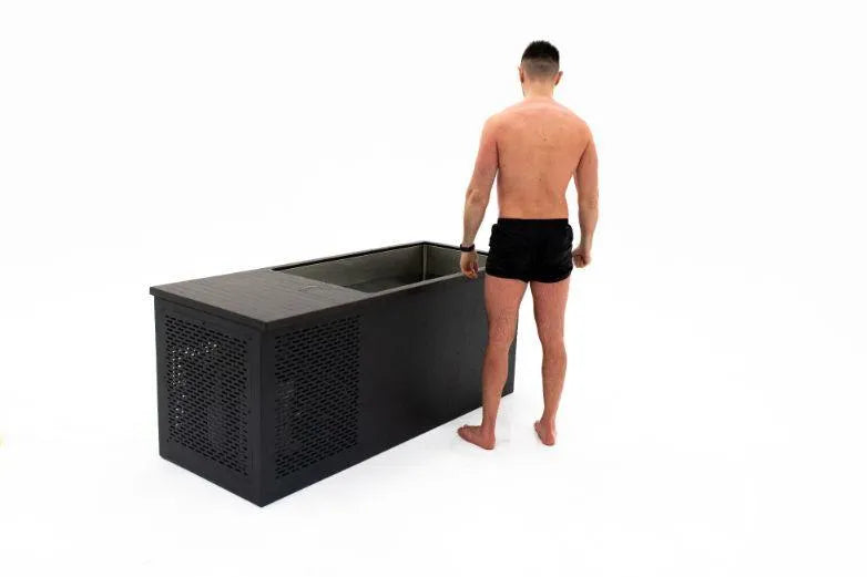 A shirtless man in black shorts stands barefoot on a white floor, facing a Chill Tubs - Professional affordable Ice Baths ideal for gym fanatics. The black rectangular tub, ideal for cold water therapy, features a perforated front panel and a partially open lid that reveals the icy interior.