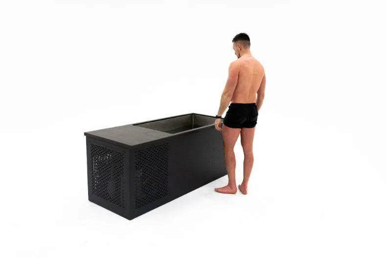 A man stands barefoot, wearing black shorts, next to a large, rectangular black container. The container appears to have a metal mesh door and an open top. He looks into the Chill Tubs - Professional affordable Ice Baths ideal for gym fanatics from Chill Tubs against a plain, white background, clearly preparing for some cold water therapy.