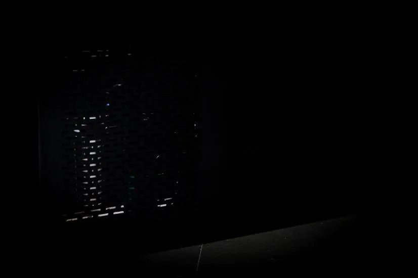 A dark, partially illuminated scene with a perforated surface barely visible on the left, casting faint light patterns. The rest of the image is shrouded in darkness, with a small section of a light gray floor showing at the bottom and a Chill Tubs - Professional affordable Ice Baths ideal for gym fanatics from Chill Tubs emerging from the shadows.